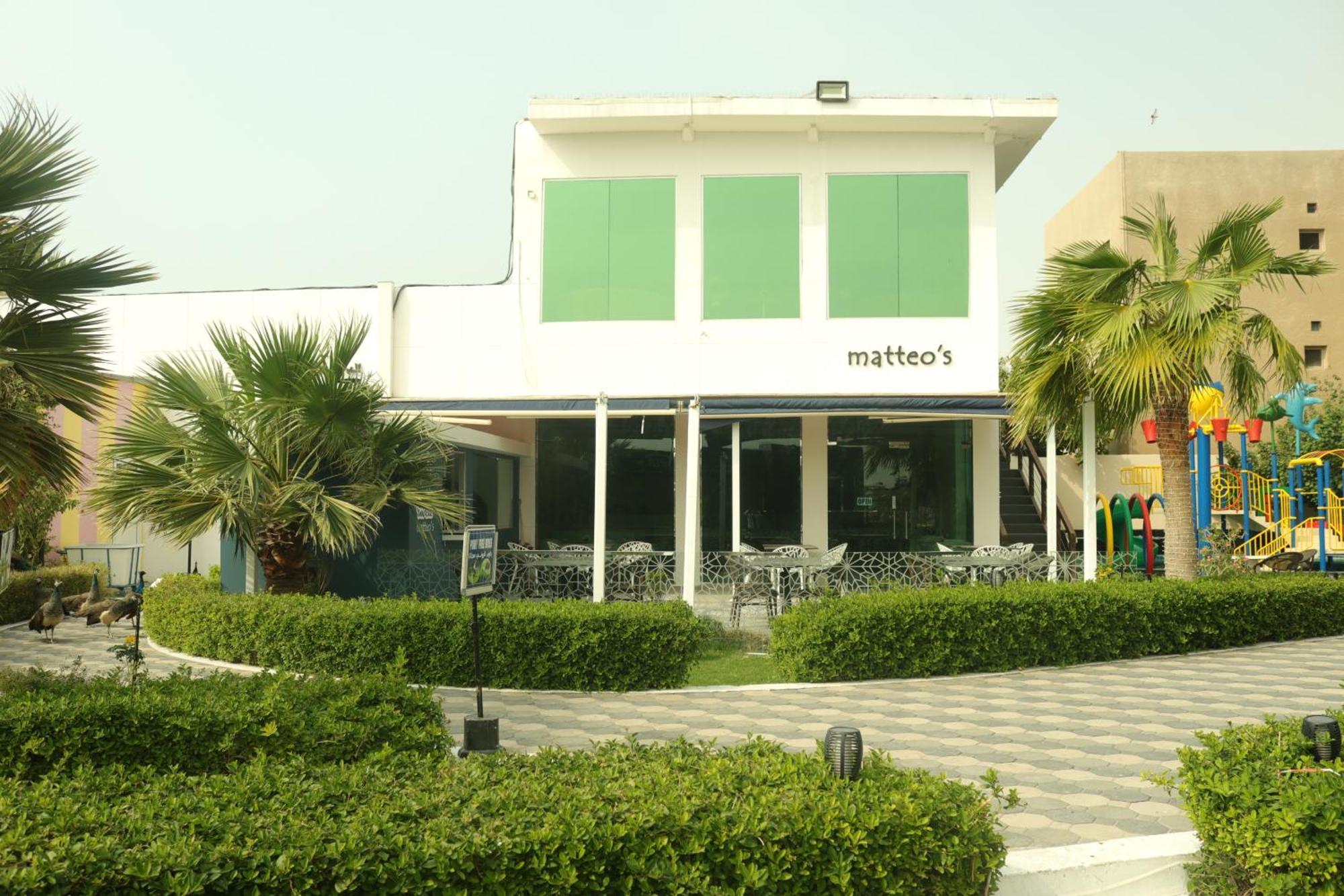 Lagoona Beach Luxury Resort And Spa Al Manamah Esterno foto The photo shows a building with the name "matteo's" displayed prominently on the front. The structure features large windows and green shutters. There is a landscaped area in front with bushes and palm trees, along with a paved pathway leading up to 