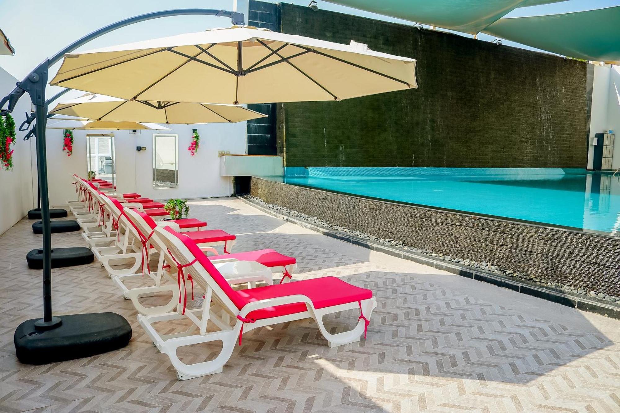 Lagoona Beach Luxury Resort And Spa Al Manamah Esterno foto The photo shows a sunny poolside area with several lounge chairs arranged in a row. Each chair features red cushions and is positioned under large, beige umbrellas for shade. In the background, there is a beautiful swimming pool with clear blue water