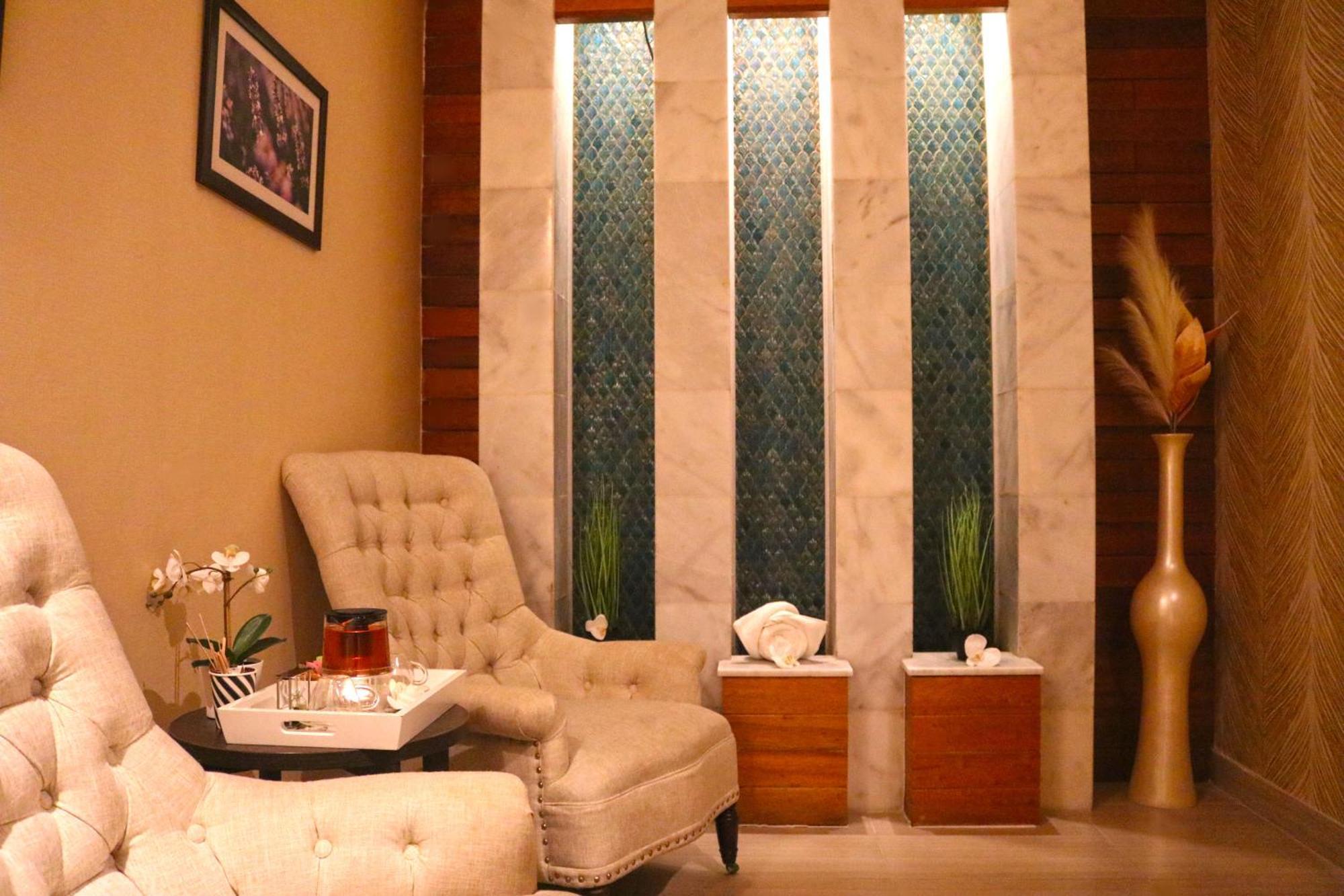 Lagoona Beach Luxury Resort And Spa Al Manamah Esterno foto The image shows a relaxing interior space, likely a waiting area or lounge. There are two plush, beige armchairs positioned near a small table, which holds a glass pitcher and a cup. The background features tall, elegant windows with decorative blue 