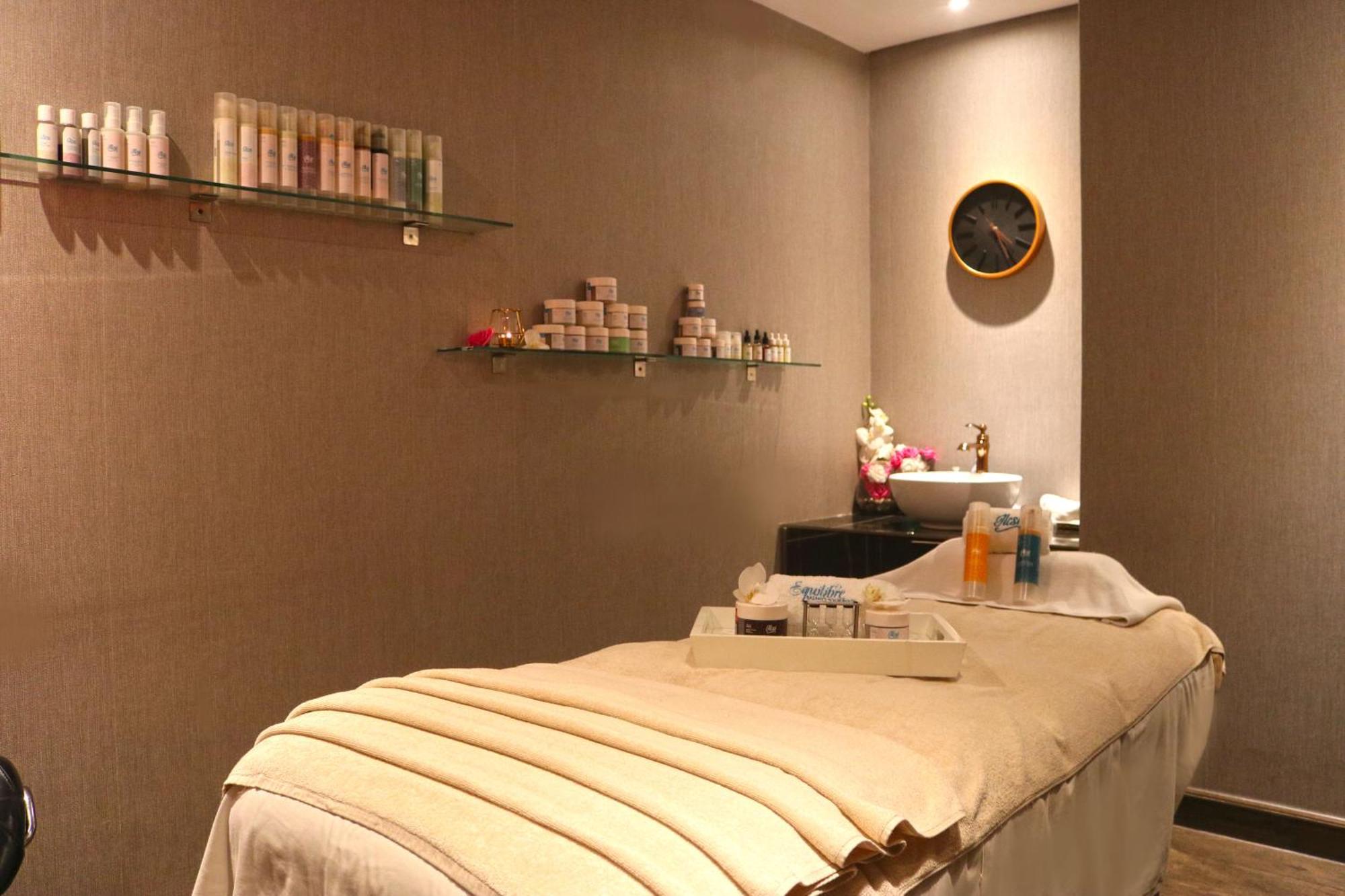 Lagoona Beach Luxury Resort And Spa Al Manamah Esterno foto The photo shows a spa treatment room. There is a massage table draped with a light-colored blanket, creating a relaxing atmosphere. On the wall, there are shelves displaying various skincare products in bottles and jars. A clock is mounted on the wal