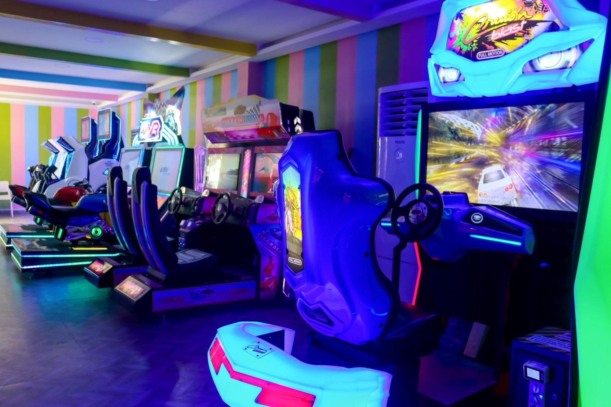 Lagoona Beach Luxury Resort And Spa Al Manamah Esterno foto The photo shows an arcade area filled with various arcade machines. The setting is illuminated with colorful neon lights, creating a vibrant atmosphere. There are several racing game machines with steering wheels and seats, each accompanied by screen