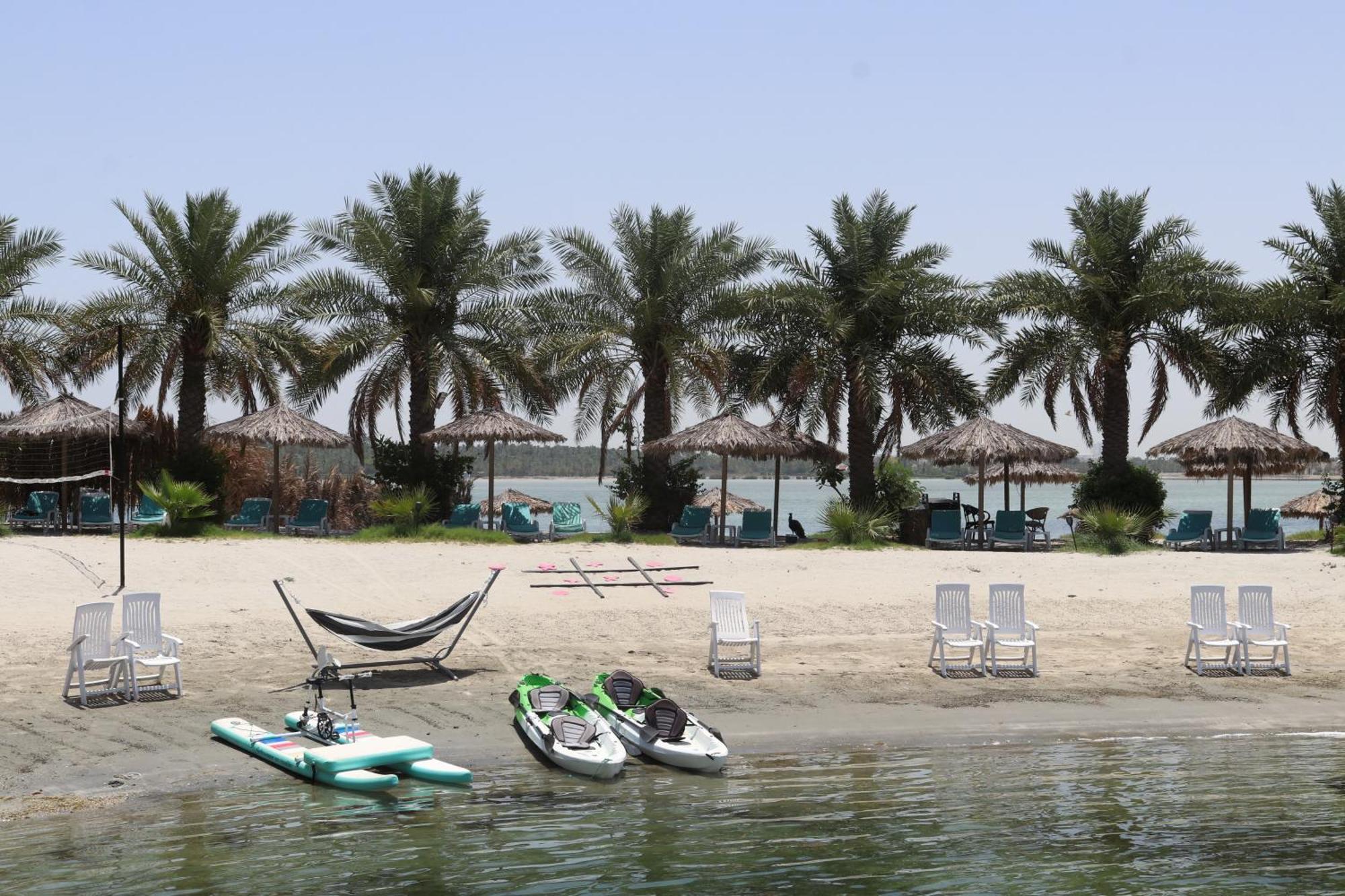 Lagoona Beach Luxury Resort And Spa Al Manamah Esterno foto The photo depicts a serene beach scene with several palm trees lining the shore. There are thatched umbrellas providing shade, and a few lounge chairs are positioned on the sand. A hammock is set up between two supports, inviting relaxation. In the f