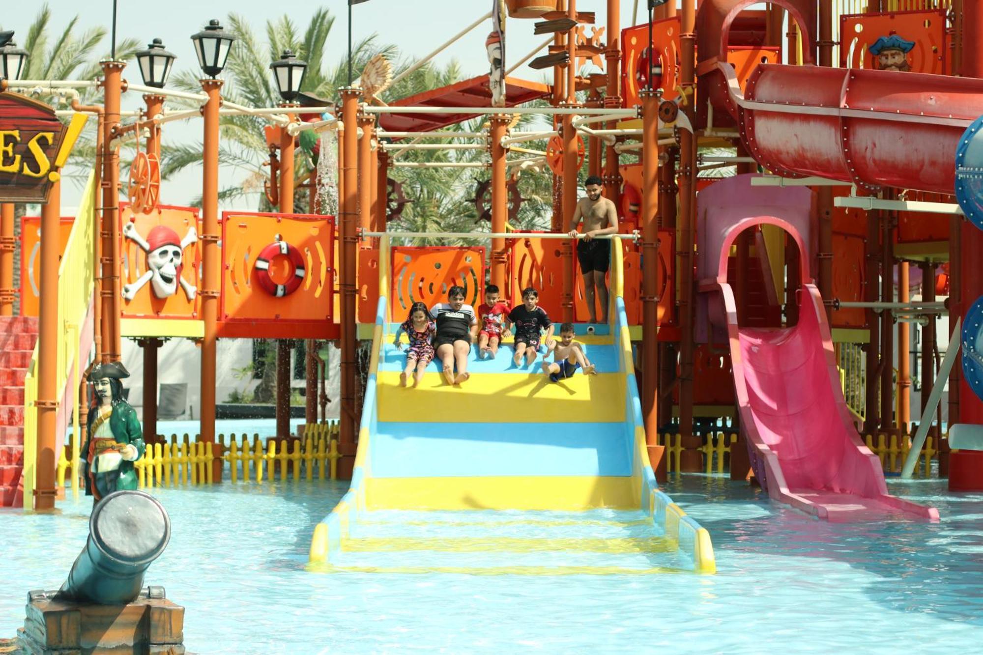 Lagoona Beach Luxury Resort And Spa Al Manamah Esterno foto The photo shows a colorful water park scene featuring a play area with multiple slides. There are children enjoying themselves, sliding down bright yellow and pink slides into a pool of water. The structure has pirate-themed decorations, including a 