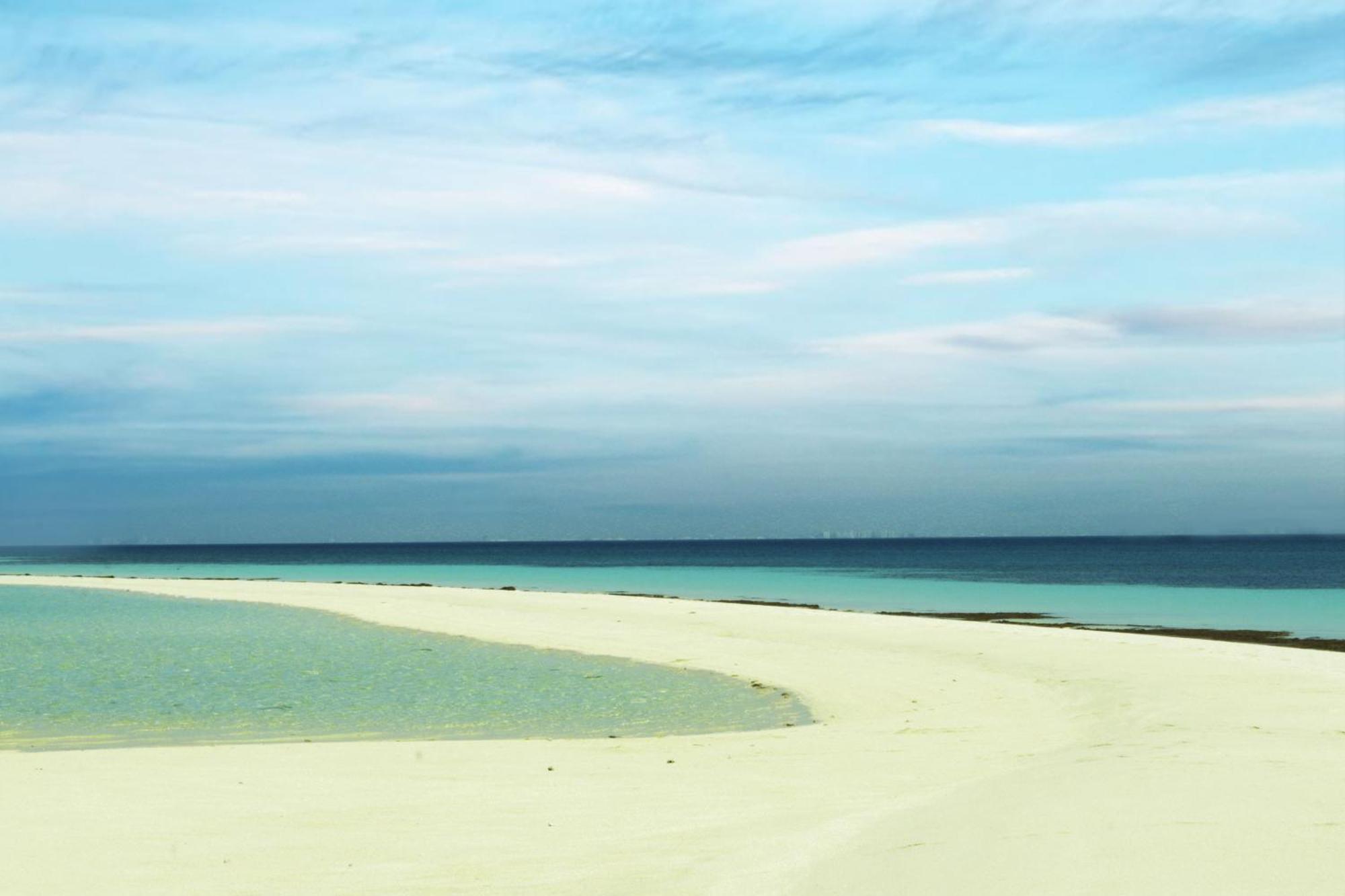 Lagoona Beach Luxury Resort And Spa Al Manamah Esterno foto The photo depicts a serene beach scene with a gentle curve of sandy shore leading into shallow turquoise waters. The calm ocean stretches into the horizon, meeting a sky filled with soft clouds in various shades of blue. The overall atmosphere is tra