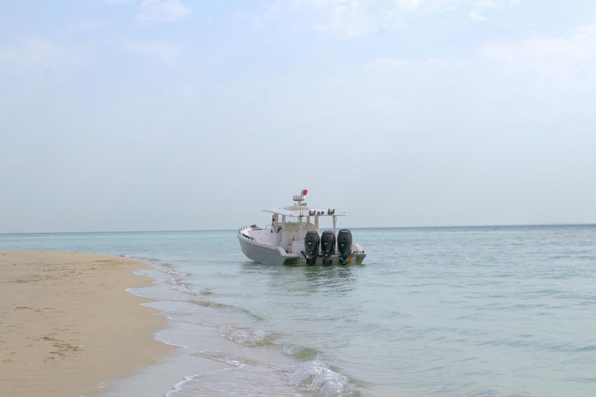 Lagoona Beach Luxury Resort And Spa Al Manamah Esterno foto The photo depicts a small boat anchored near a sandy beach, partially submerged in calm, clear water. The boat is white and features three outboard motors on its stern. In the background, the sea stretches out toward the horizon under a light, cloudy