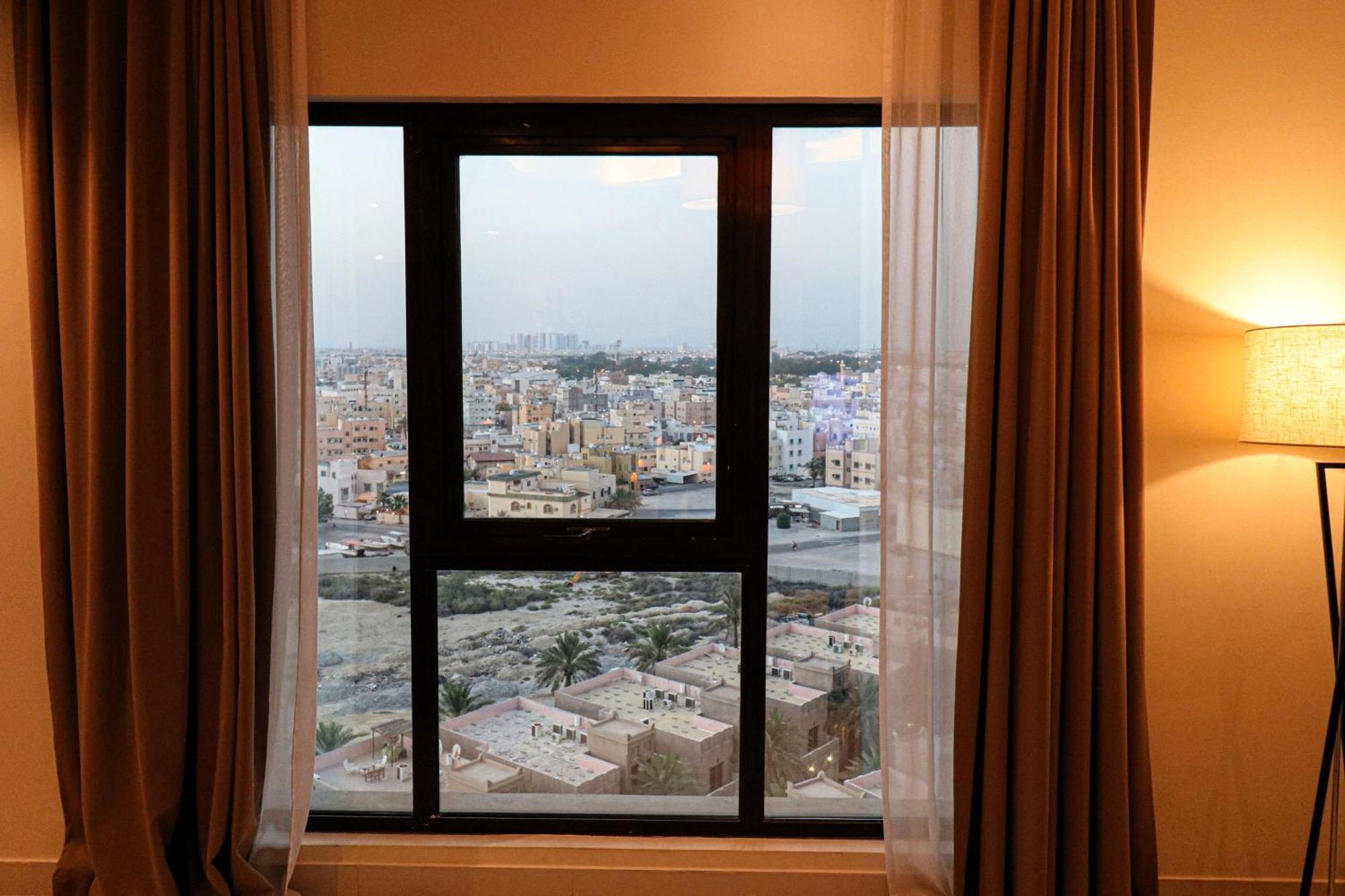 Lagoona Beach Luxury Resort And Spa Al Manamah Esterno foto The photo shows a window with a black frame and sheer curtains. Through the window, there is a view of a cityscape, displaying a densely populated area with various buildings and rooftops. The sky appears slightly overcast, suggesting a muted lightin