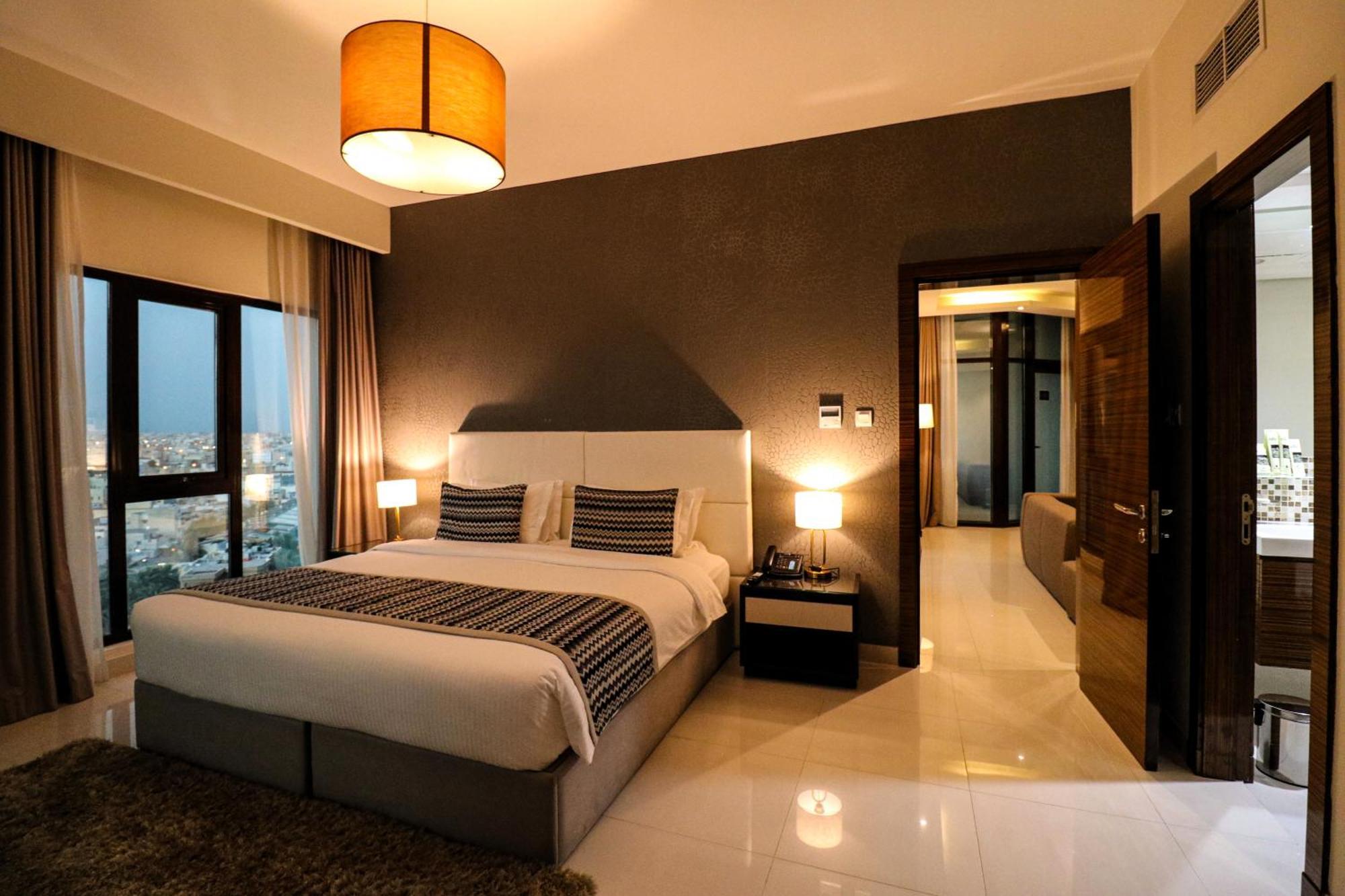 Lagoona Beach Luxury Resort And Spa Al Manamah Esterno foto The photo depicts a modern hotel room or suite. It features a large bed with a white and gray color scheme, accented with decorative pillows. On either side of the bed, there are sleek bedside lamps providing warm lighting. The walls are a dark color