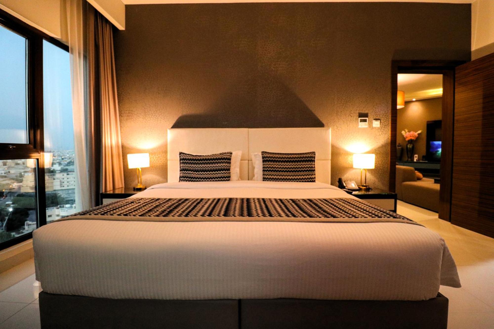 Lagoona Beach Luxury Resort And Spa Al Manamah Esterno foto The photo depicts a modern hotel room featuring a large bed with a white comforter and two decorative pillows. The headboard is light-colored, and the walls are painted a dark hue. On either side of the bed, there are bedside tables with lamps. The r