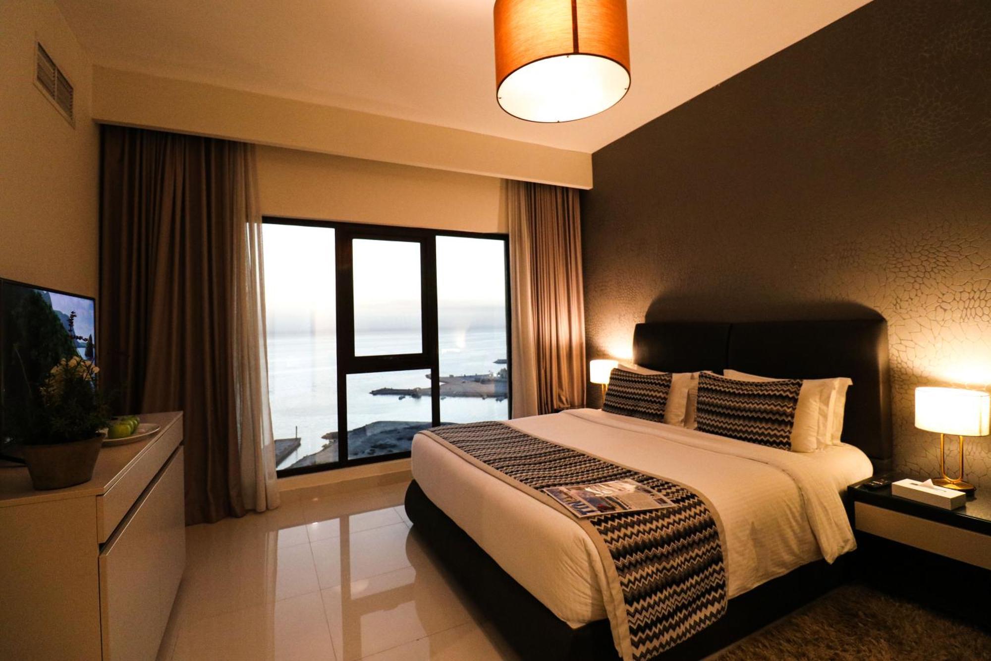Lagoona Beach Luxury Resort And Spa Al Manamah Esterno foto The photo shows a modern hotel room featuring a neatly made bed with decorative pillows. The room has a stylish design with a dark accent wall and a large window that offers a view of the outdoors. Natural light comes in, giving the space a bright an