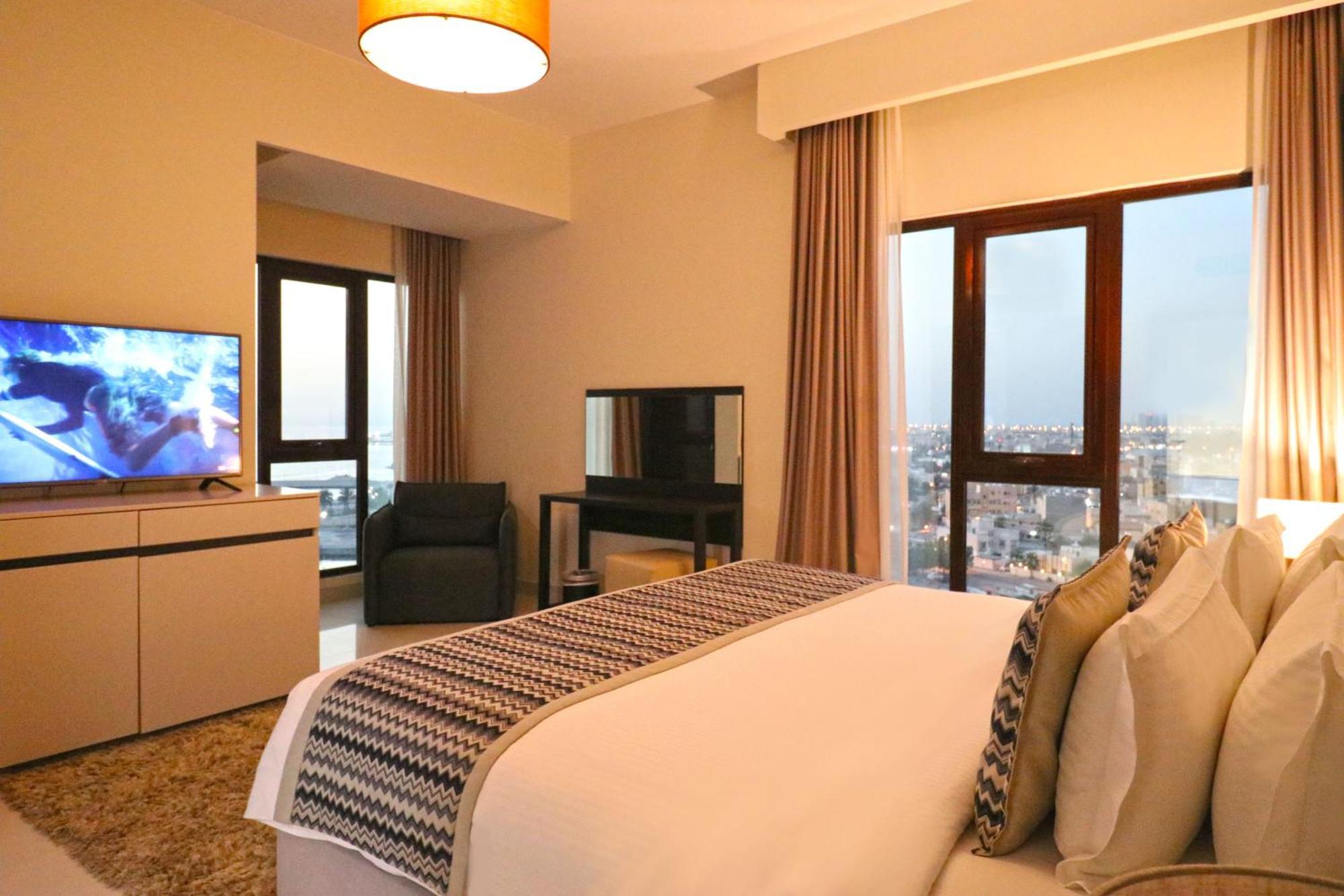 Lagoona Beach Luxury Resort And Spa Al Manamah Esterno foto The photo shows a modern hotel room or suite. It features a neatly made bed with decorative pillows in the foreground. To the left, there is a TV on a cabinet, and further back, a comfortable chair is situated in front of a large window that likely o