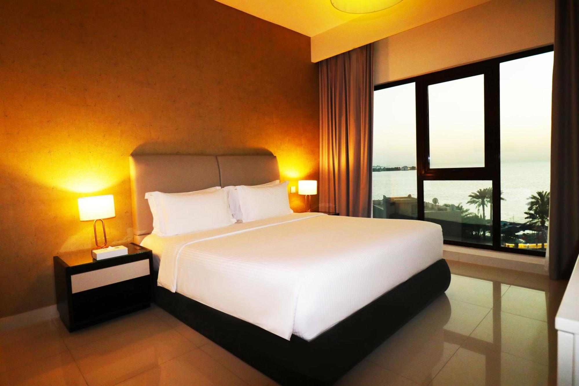 Lagoona Beach Luxury Resort And Spa Al Manamah Esterno foto The photo shows a modern hotel bedroom with a large, comfortable bed dressed in white linens. The room features warm lighting, including bedside lamps, creating a cozy atmosphere. There are large windows with a view of the outdoors, possibly overlook