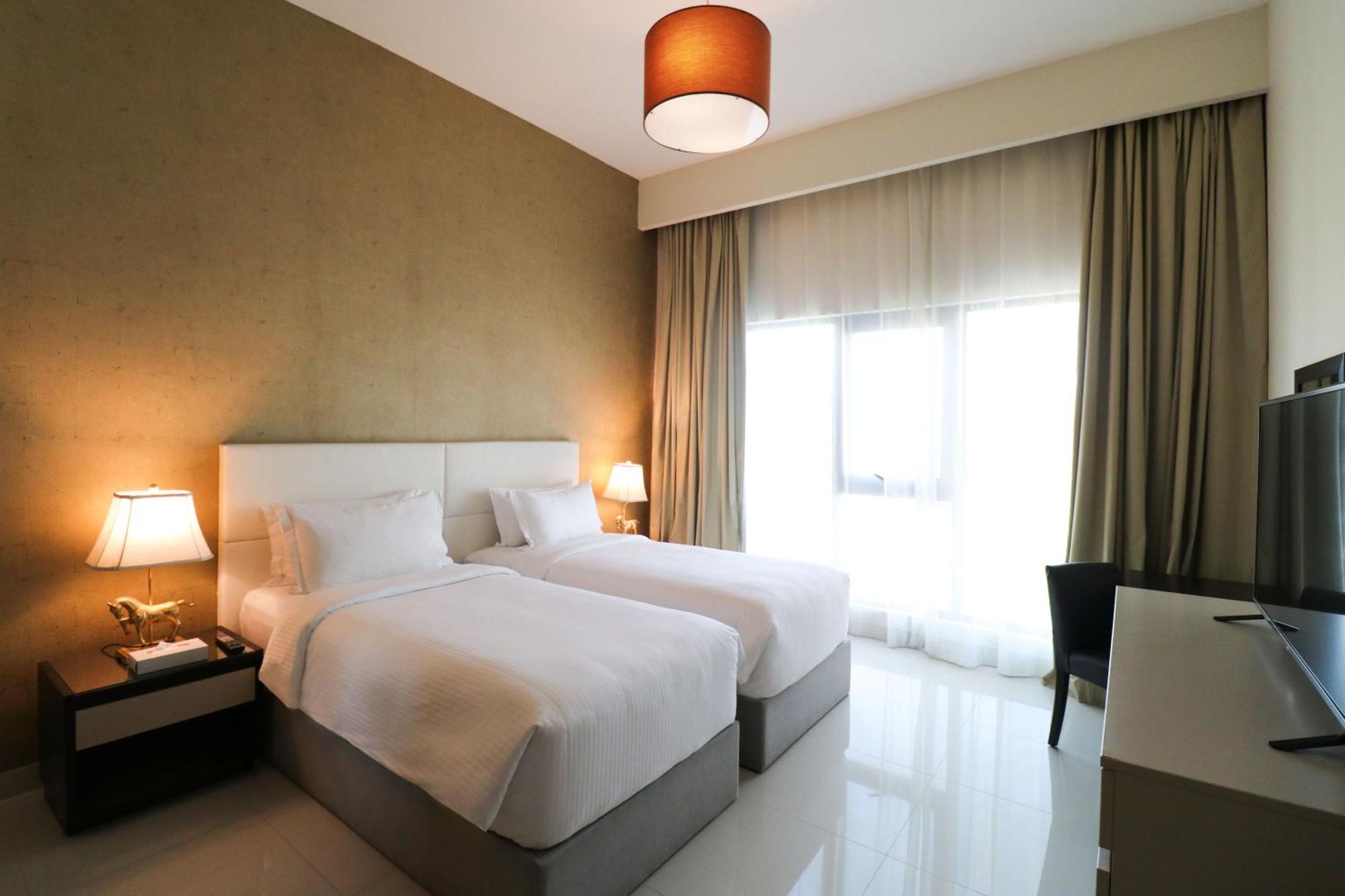 Lagoona Beach Luxury Resort And Spa Al Manamah Esterno foto The photo shows a hotel room featuring two single beds with white bedding. The walls have a neutral tone, and there's a large window covered by light, sheer curtains allowing natural light to enter. On either side of the beds, there are bedside table