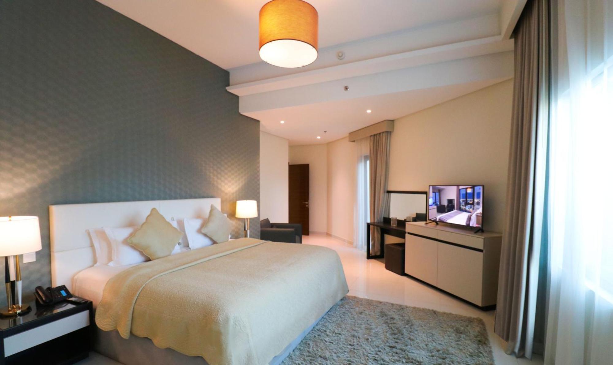 Lagoona Beach Luxury Resort And Spa Al Manamah Esterno foto The photo shows a modern hotel room featuring a large bed with a light-colored blanket and decorative pillows. The walls are painted in a subtle shade, and there is a textured accent wall behind the bed. On one side, a bedside lamp provides warm ligh