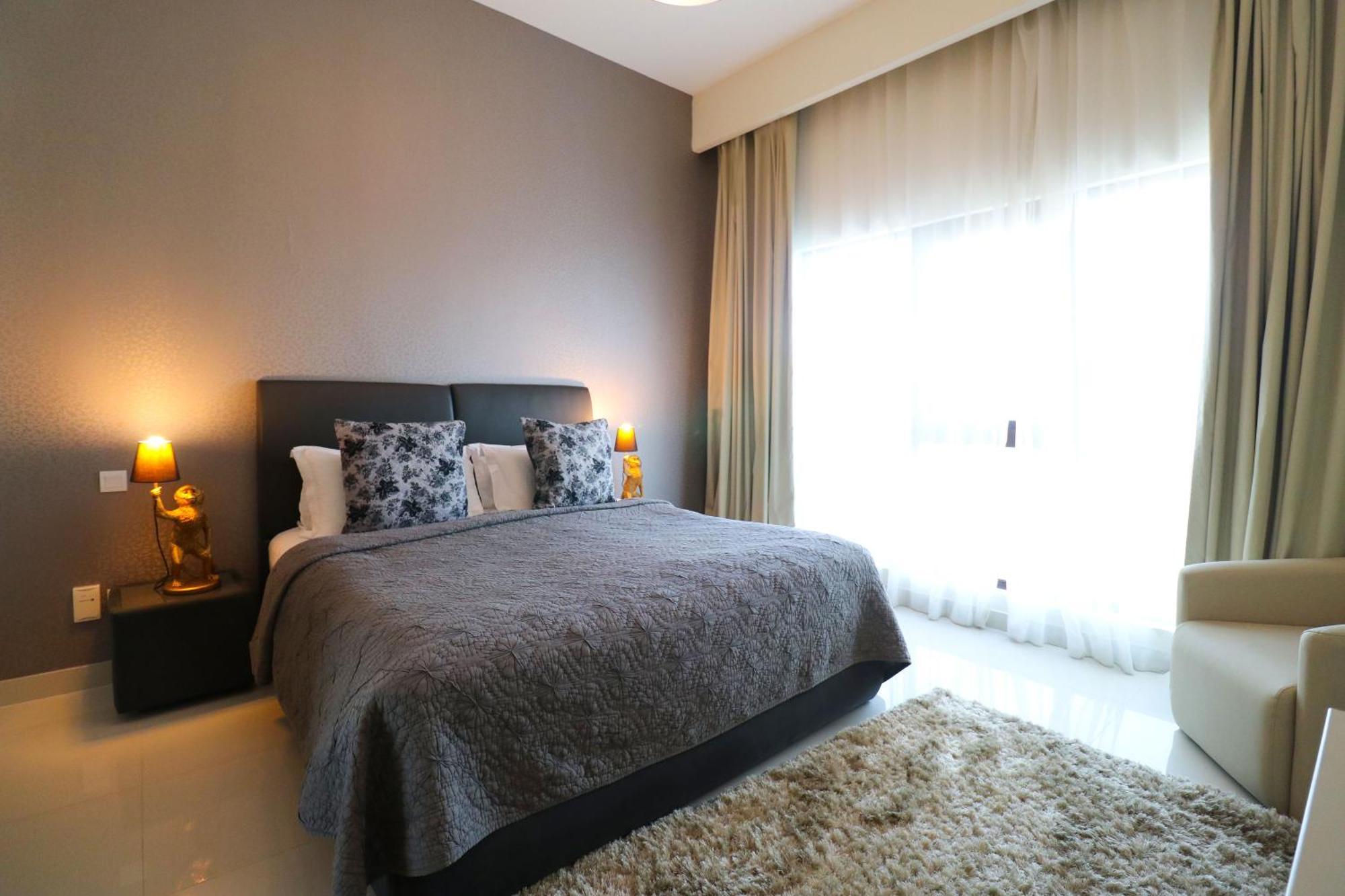 Lagoona Beach Luxury Resort And Spa Al Manamah Esterno foto The photo shows a stylish bedroom with a modern design. It features a large bed with a dark gray bedspread and decorative pillows. On either side of the bed are two wooden nightstands, each with a lamp. There is a large window covered with light, flo
